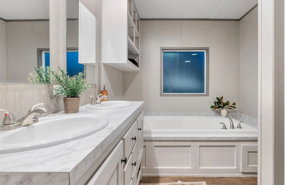 Modern bathroom with double sinks, a marble countertop, a built-in bathtub, a window, and potted plants on the counter and tub ledge.