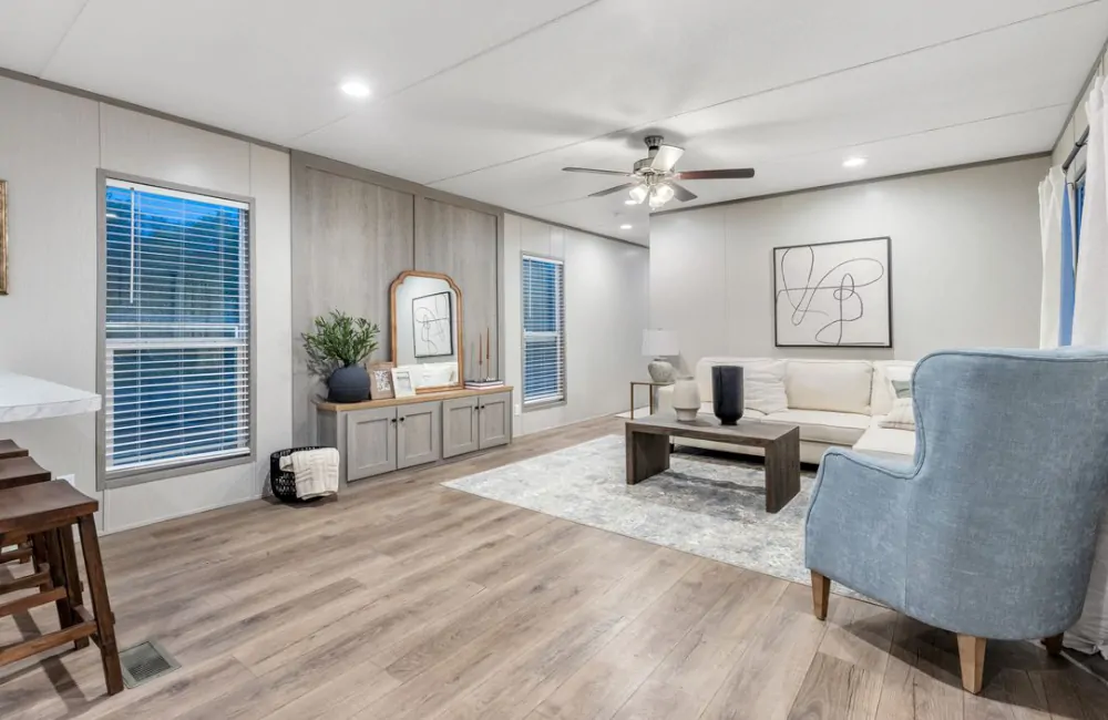 Modern living room with wood flooring, neutral walls, a white sectional sofa, blue armchair, coffee table, large windows, and minimalist decor.