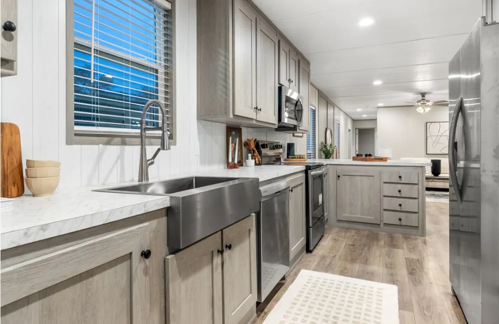 Modern kitchen with stainless steel appliances, farmhouse sink, gray cabinets, marble countertops, and wood flooring, viewed from the sink toward an open living area.