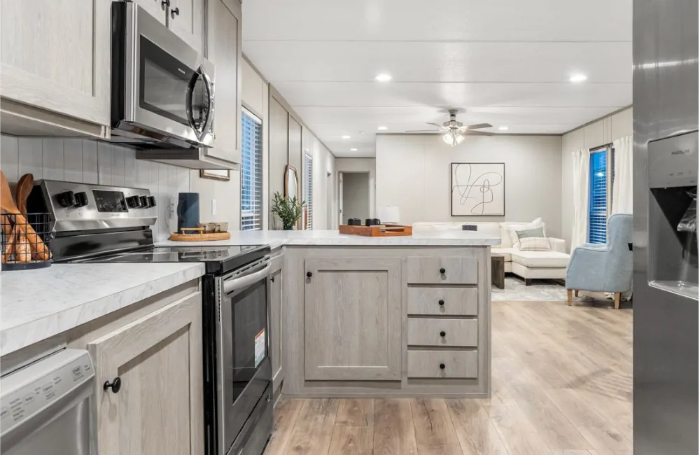 Modern kitchen with light wood cabinets, stainless steel appliances, marble countertops, and an open view into a furnished living room with a ceiling fan.