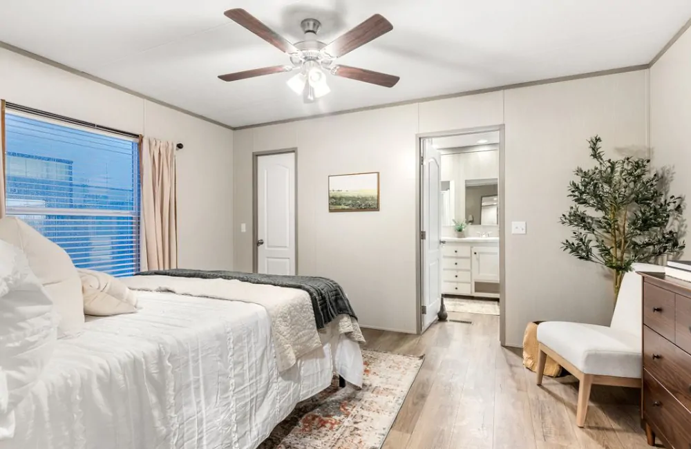 A bedroom with a neatly made bed, a ceiling fan, a dresser, a chair, and an open door leading to a well-lit bathroom with a vanity and mirror.