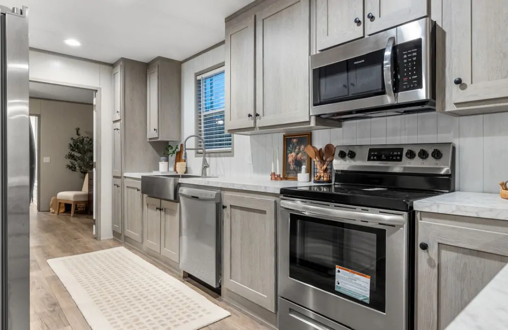 Modern kitchen with light gray cabinets, stainless steel appliances, farmhouse sink, marble countertops, and a beige runner rug on wooden flooring.
