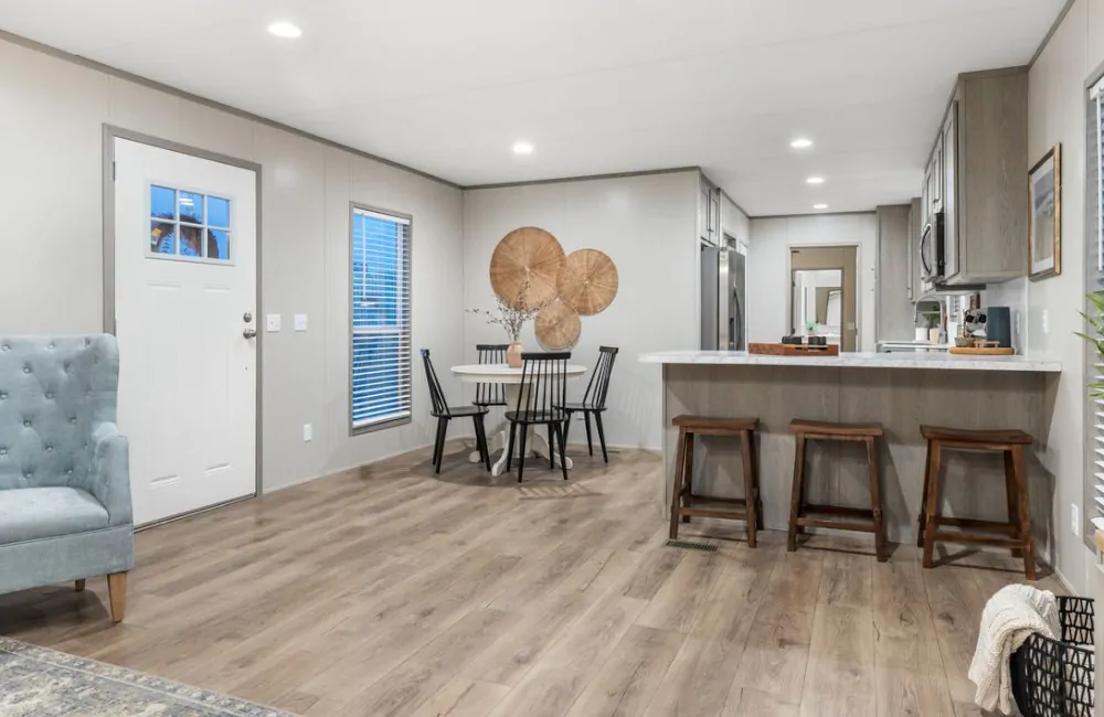 Open-plan living space with wood flooring, featuring a small dining table, kitchen with island and stools, light walls, and minimal decor.