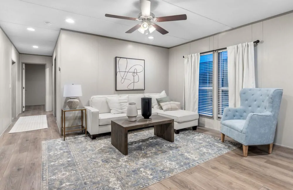 Modern living room with a white sectional sofa, blue tufted armchair, wooden coffee table, abstract wall art, and large windows with white curtains on a patterned rug.