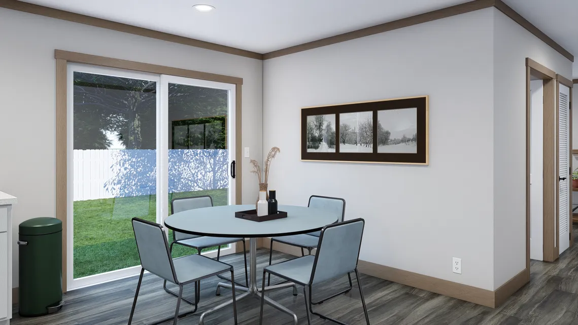 A small dining area with a round table and four chairs is next to sliding glass doors overlooking a backyard. A framed picture hangs on the wall above gray wood flooring.