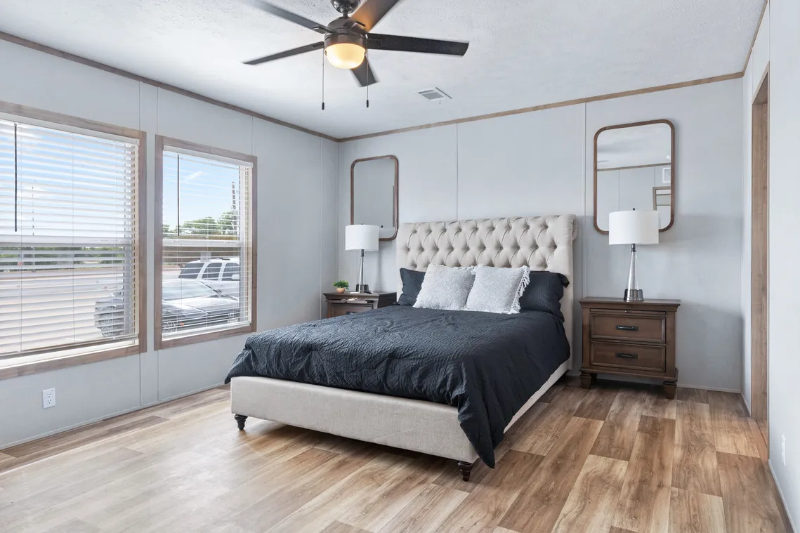 A bedroom with a bed featuring a tufted headboard, dark bedding, two bedside tables with lamps, large windows, wood flooring, and a ceiling fan.