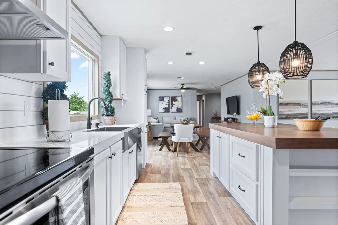 Modern kitchen with white cabinets, wood countertops, pendant lights, and a view into a dining and living area; large window provides natural light.