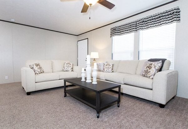 A living room with two beige sofas, decorative pillows, a black coffee table with white vases, a table lamp, and two windows with patterned valances.