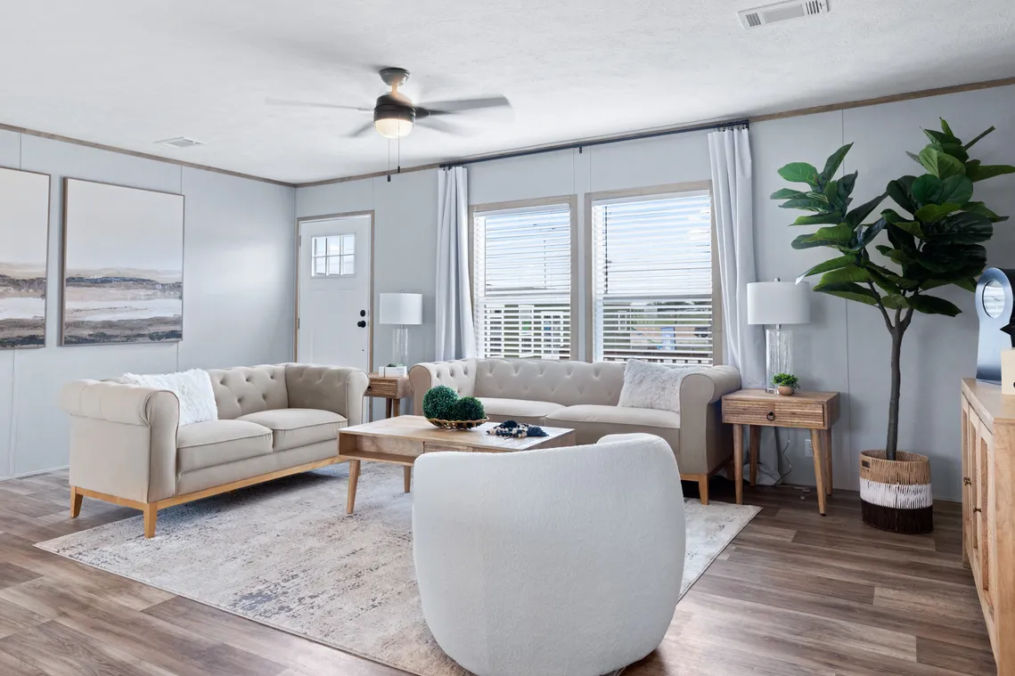 Modern living room with light-colored sofas, a round chair, wooden coffee and side tables, large windows with white curtains, wall art, and a potted plant on wood flooring.
