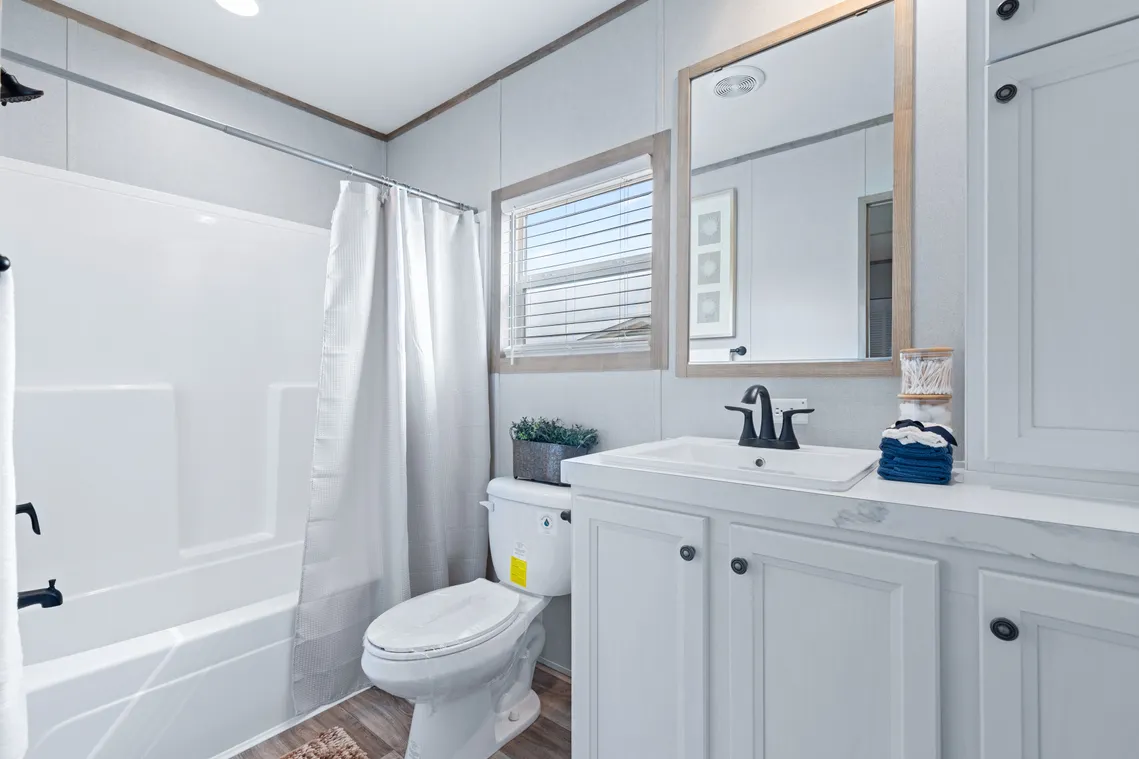 Modern bathroom with white cabinets, a sink with black fixtures, a toilet, a bathtub with a shower curtain, and a window above the toilet.