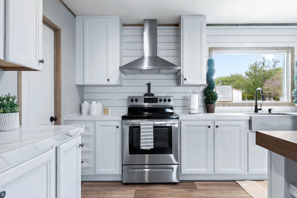 Modern kitchen with white cabinets, stainless steel stove and hood, large window above sink, and wood flooring. Potted plants and kitchen essentials are arranged on the counters.