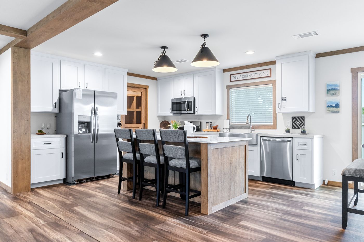 Modern kitchen with stainless steel appliances, white cabinets, a central island with four gray chairs, wood accents, and pendant lights, featuring a sign above the window.