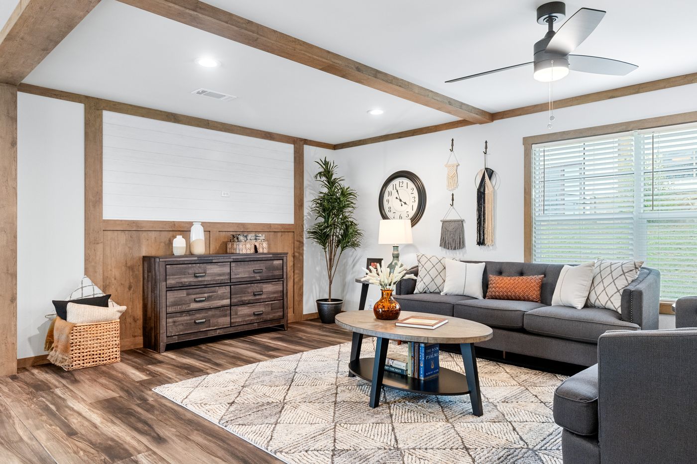 Modern living room with gray sofa, patterned rug, wooden coffee table, dresser, wall clock, decorative wall hangings, ceiling fan, large window, and potted plant on wood-look floors.