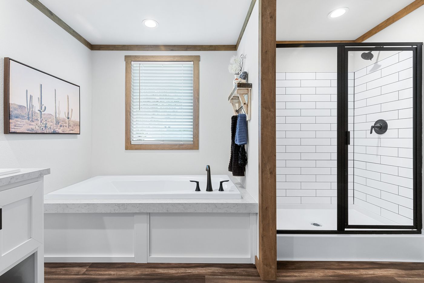 Modern bathroom with a white bathtub, black fixtures, a glass-enclosed shower, wooden accents, and a window with blinds. A cactus artwork hangs on the wall.