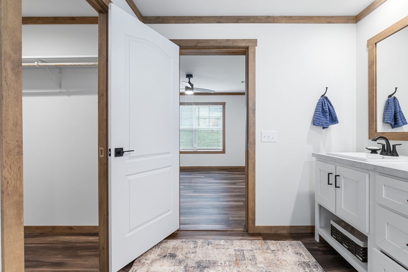 A bathroom with white walls, a vanity with a sink and mirror, a door leading to a walk-in closet, and a view into a room with large window and wood flooring.