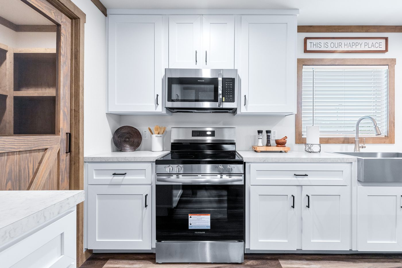 Modern kitchen with white cabinets, stainless steel oven and microwave, farmhouse sink, and a window with blinds. Sign above window reads "This is our happy place.