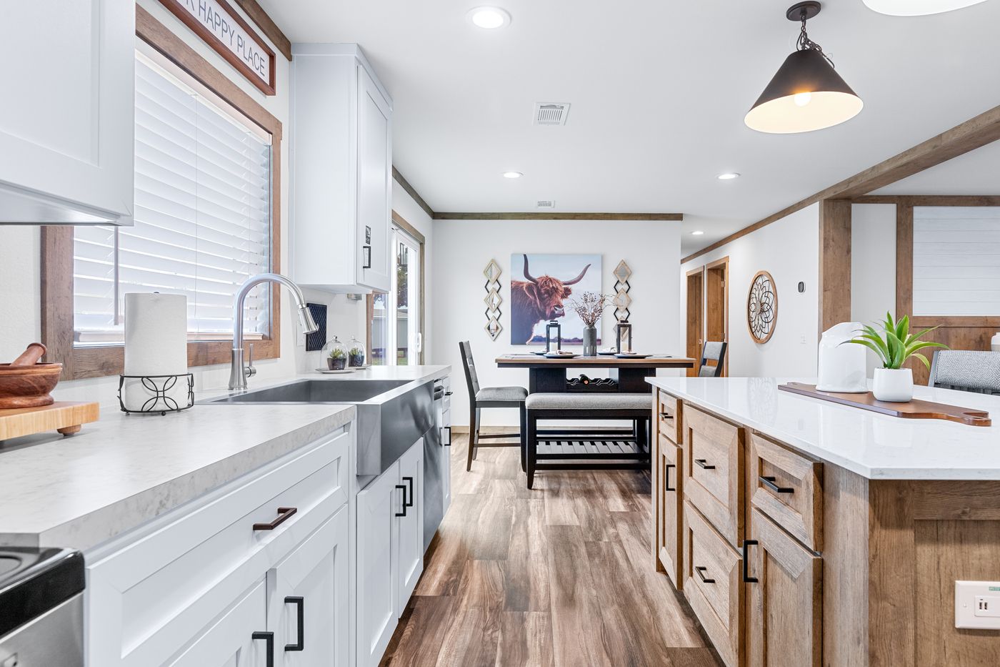 Modern kitchen with white cabinets, wooden accents, an island with stools, stainless steel sink, and wall art featuring a bull and decorative plates in the dining area.