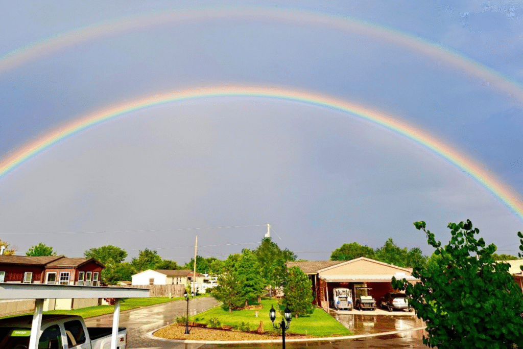 Two bright rainbows arch across a cloudy sky over a residential area with houses, green trees, and wet pavement after rain.