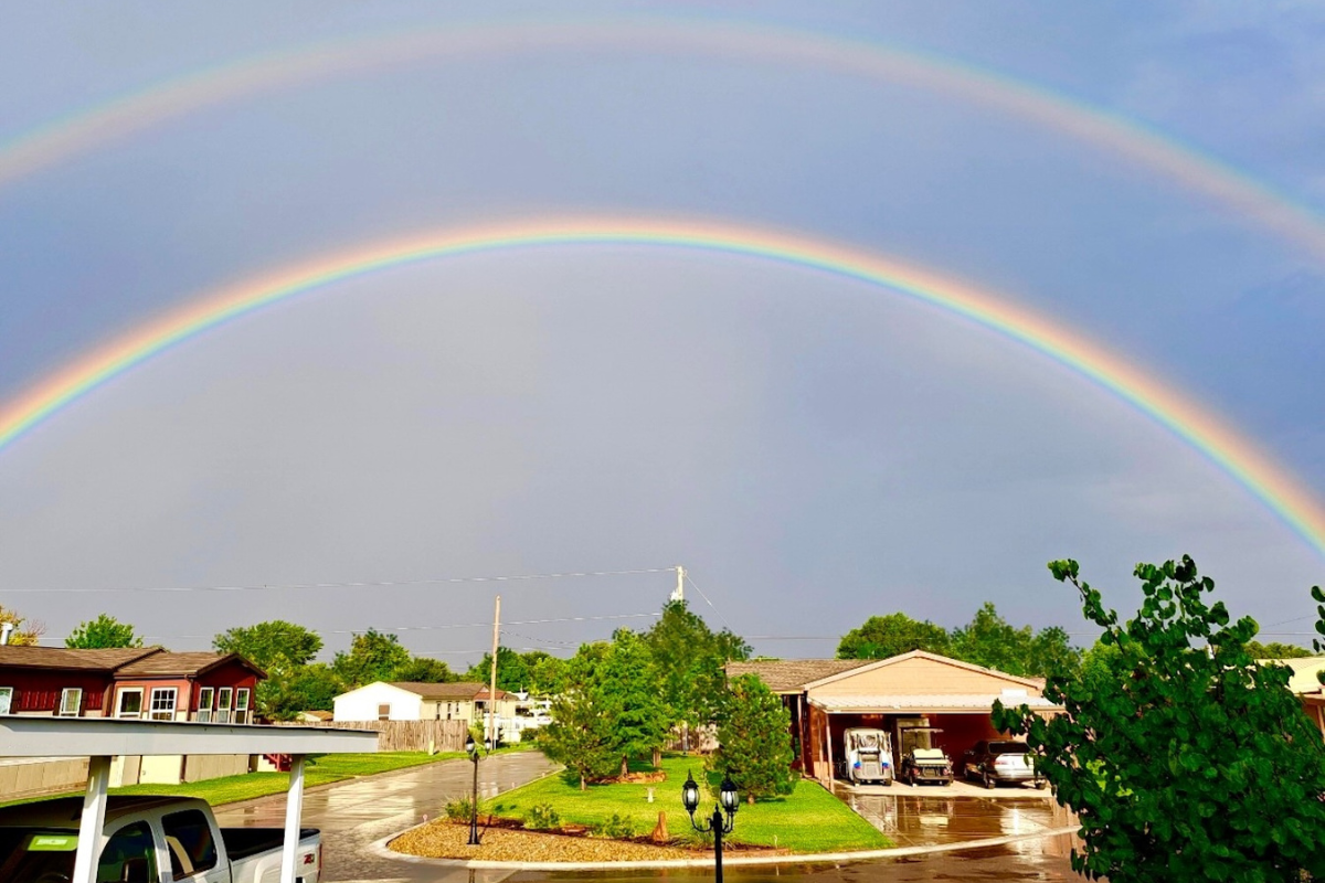 Two bright rainbows arch across a cloudy sky over a residential area with houses, green trees, and wet pavement after rain.