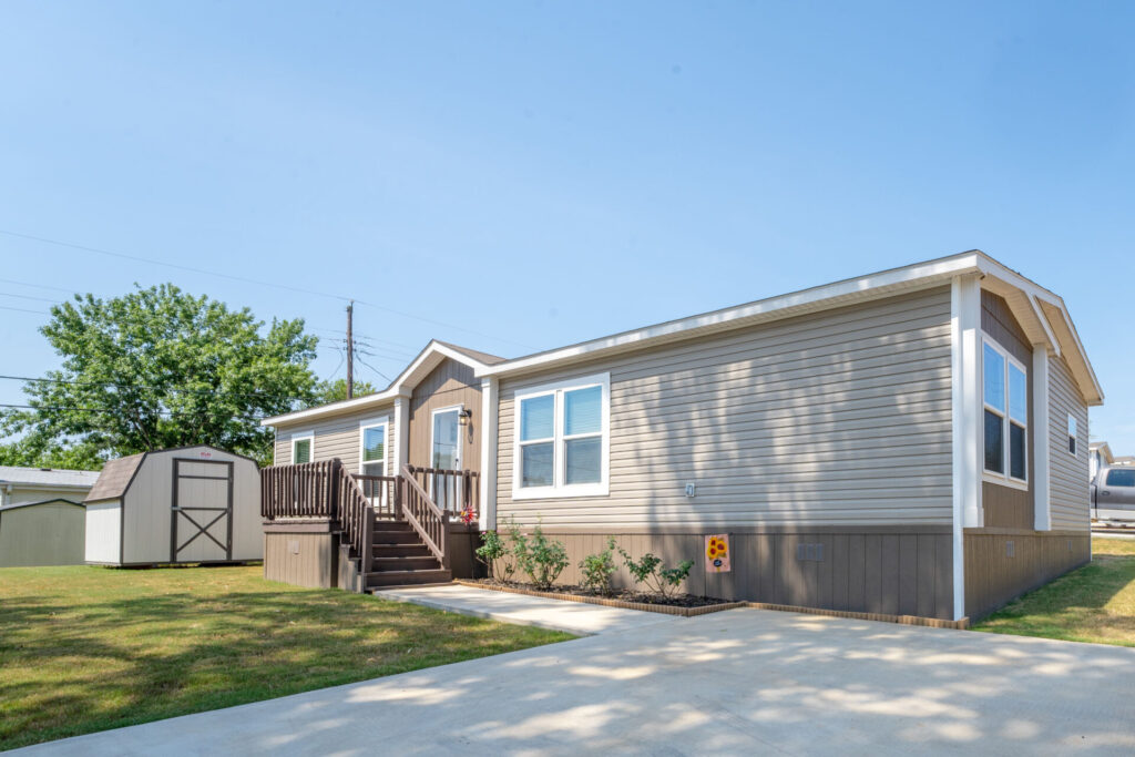 A beige manufactured home with white trim sits on a concrete driveway with a small shed and green lawn in the background under a clear blue sky.