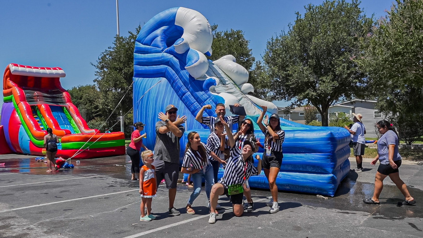 A group of people, some in striped shirts, pose in front of large inflatable water slides in a parking lot on a sunny day. Other individuals are visible in the background.