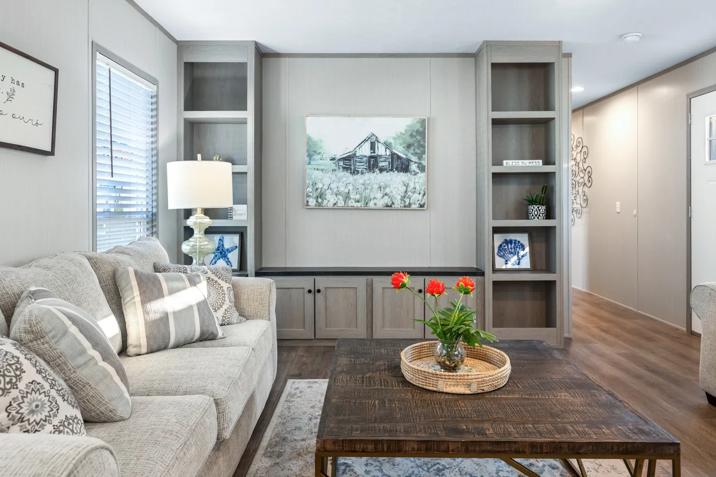 A modern living room with a beige sofa, wooden coffee table, potted plant, built-in shelves, and a painting of a barn on the wall.
