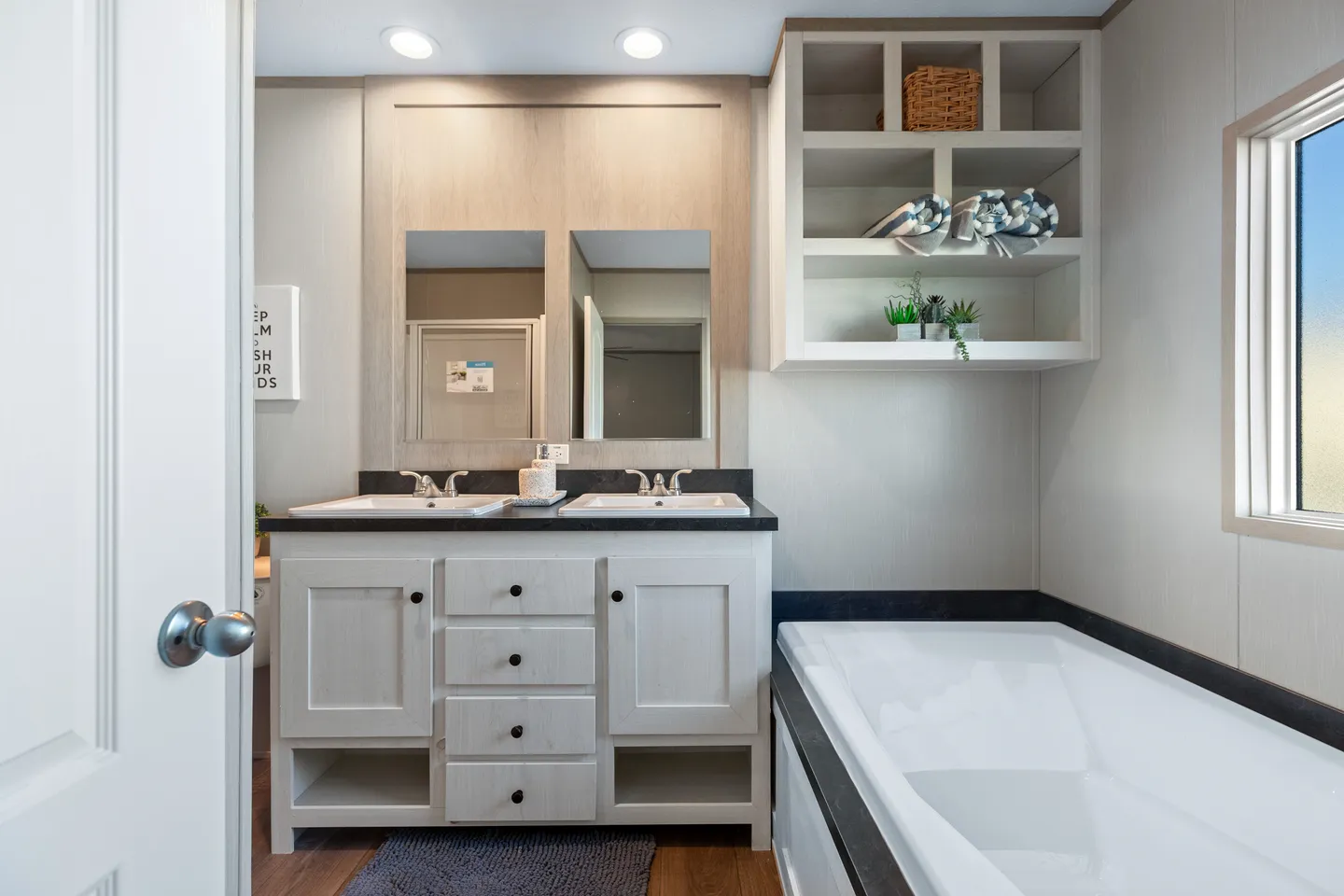 Modern bathroom with double sinks, a built-in bathtub, open shelves with towels and decor, large mirrors, and recessed lighting.