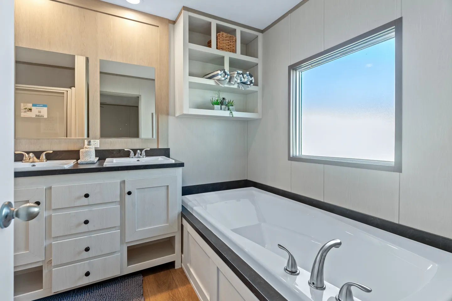 Modern bathroom with a large built-in bathtub, double sinks with mirrors, a window, and open shelves with decorative items above the tub.