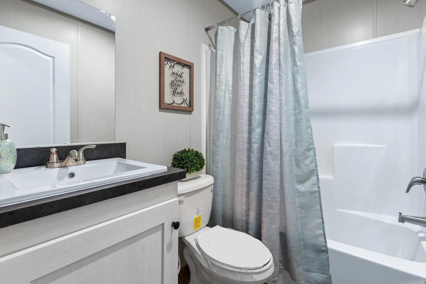 A small bathroom with a white sink, black countertop, toilet, shower with a white tub and gray curtain, and a framed sign above the toilet.