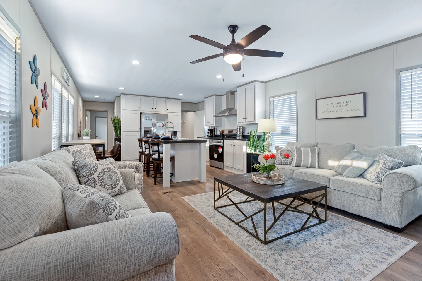Modern open-plan living room and kitchen with neutral decor, two sofas, a coffee table with flowers, dining area, and ceiling fan. Natural light enters through multiple windows.