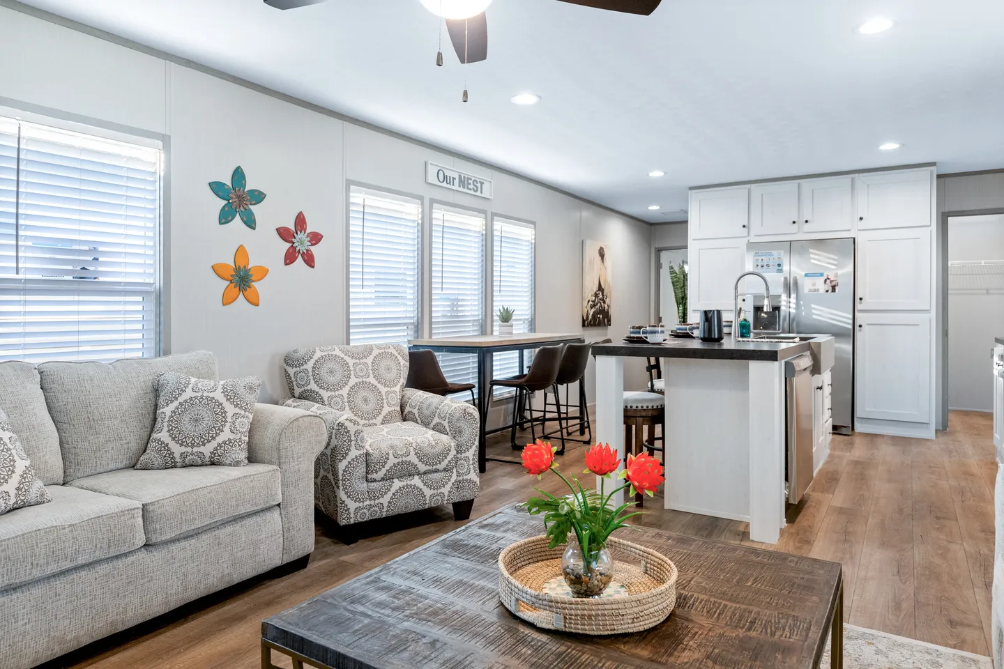 Modern open-concept living room and kitchen with neutral décor, patterned furniture, a wooden coffee table with red flowers, and wall art featuring colorful flowers and birds.