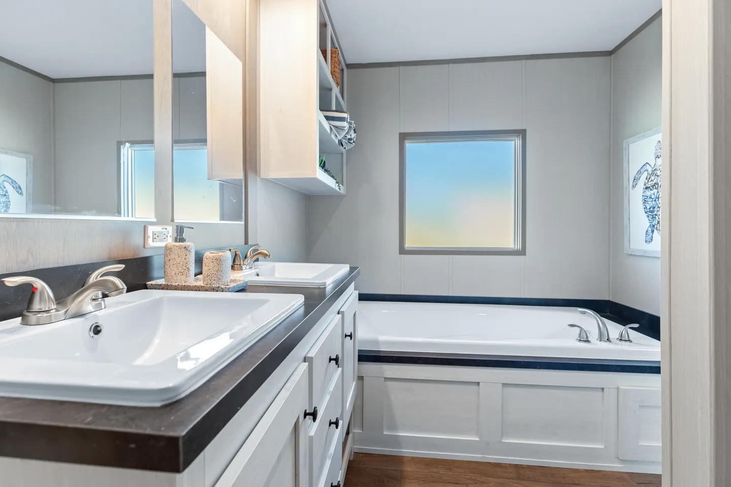 Modern bathroom with double sinks, a built-in bathtub, two windows, and light-colored walls; turtle artwork is visible on the wall near the tub.
