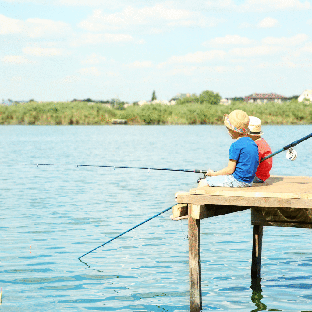 Two children wearing hats sit on a wooden dock, fishing with rods over a calm lake, with reeds and houses visible in the background.