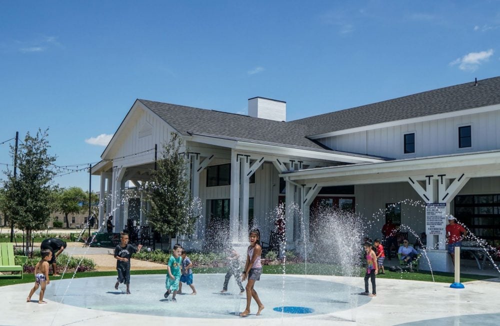 Children play in a splash pad fountain in front of a modern white building on a sunny day.