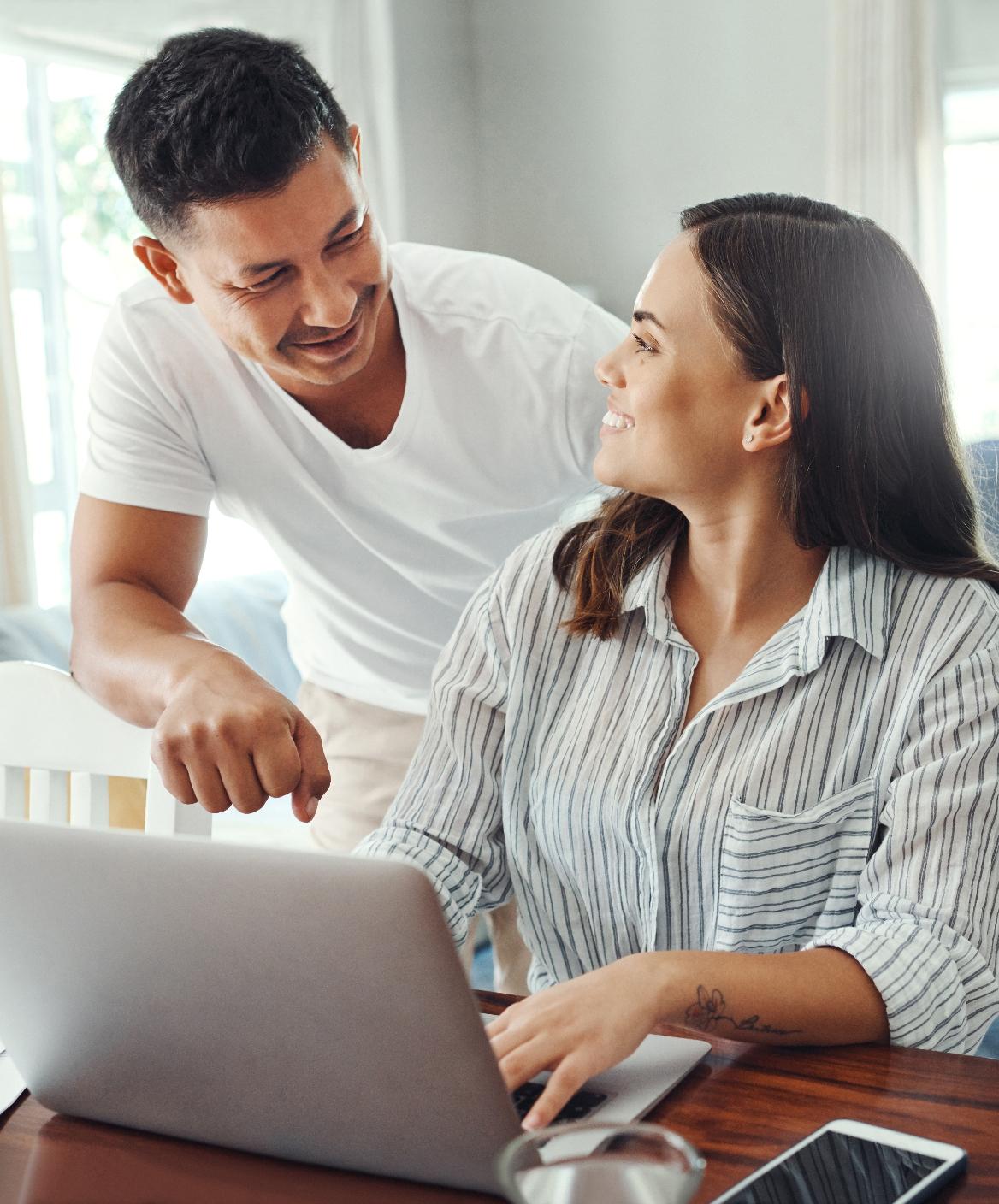 A man stands beside a woman who is seated at a table working on a laptop. They are both smiling and looking at each other in a bright room.