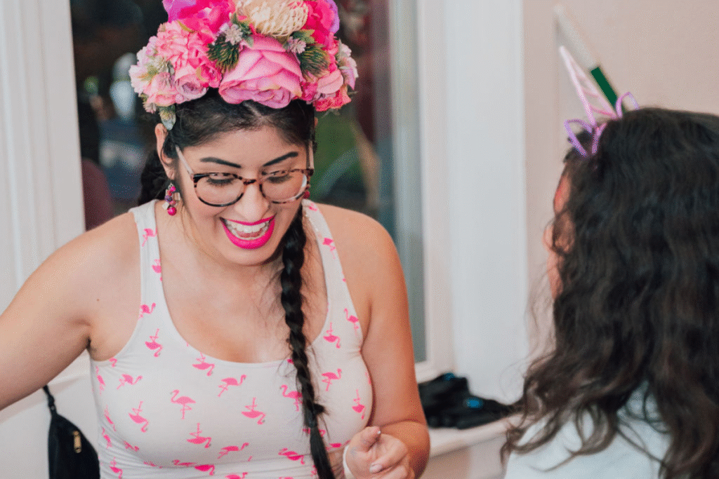 A woman wearing glasses, a flower crown, and a flamingo-patterned tank top smiles and talks to another person in a unicorn headband.