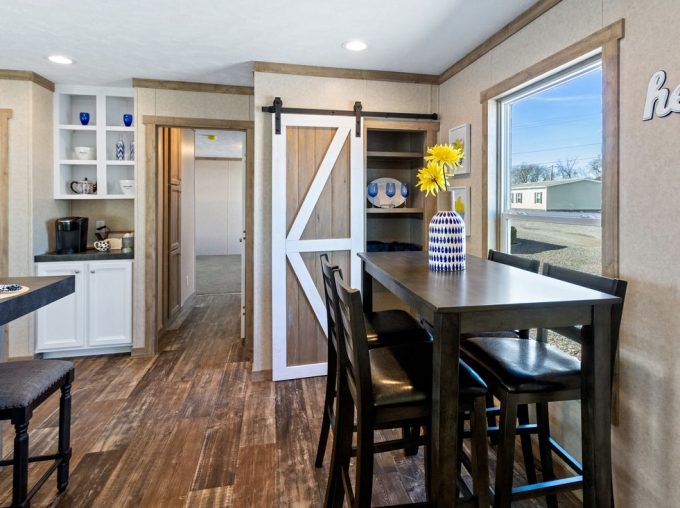 A dining area with a tall dark table, four chairs, a vase of yellow flowers, a sliding barn door, wood flooring, and shelves with dishes in a bright, modern interior.