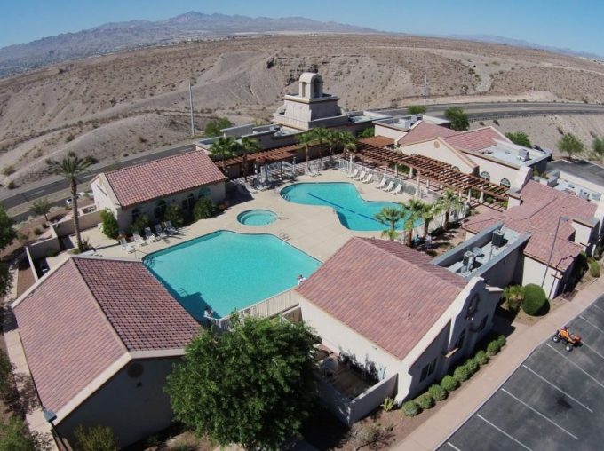 Aerial view of a large outdoor swimming pool surrounded by lounge chairs and buildings with red-tiled roofs, set in a desert landscape near a road.