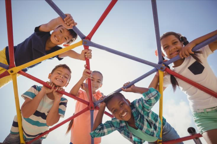 Five children are smiling and playing on a colorful jungle gym, viewed from below against a bright, sunny sky.