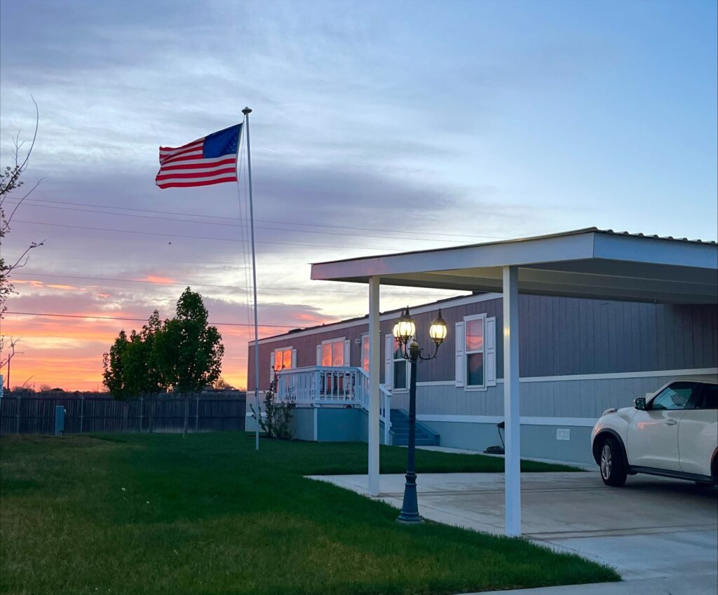 A white car is parked in the driveway outside a gray manufactured home with a covered porch. An American flag flies on a pole beside the house at sunset.