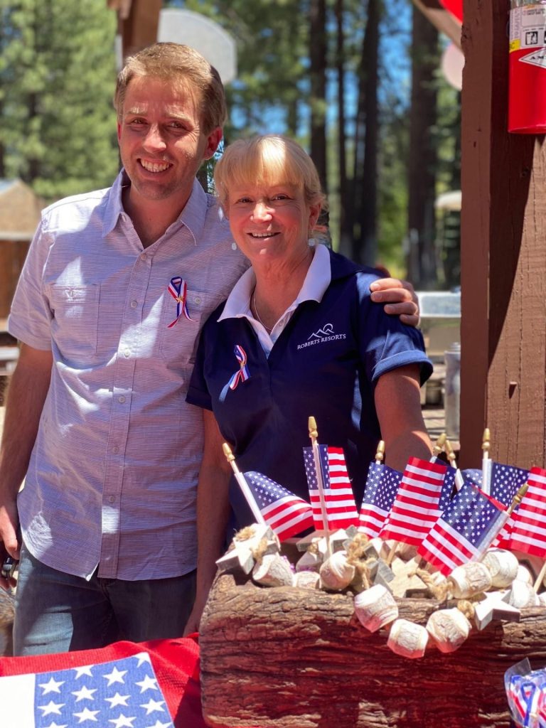 A man and a woman stand outdoors by a table decorated with small American flags and patriotic ribbons, surrounded by trees and sunlight.