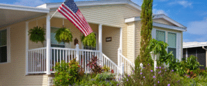 A yellow manufactured home with a white porch, potted plants, hanging ferns, and an American flag, surrounded by a landscaped garden on a sunny day.