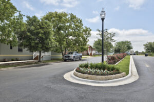 A quiet residential street with landscaped median, trees, parked pickup truck, and manufactured homes on a sunny day.