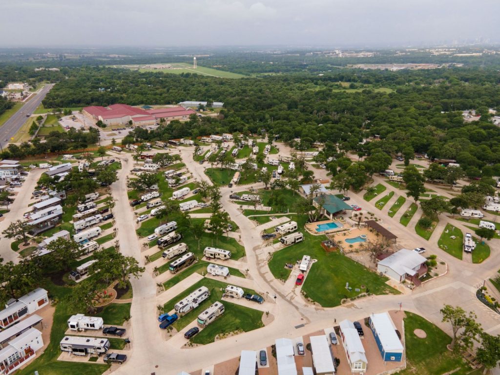 Aerial view of an RV park with numerous trailers, swimming pool, small buildings, and surrounding green trees under a cloudy sky.