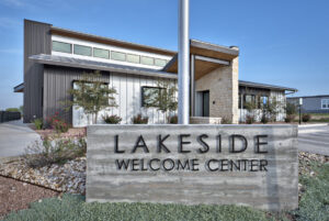 Modern building with large windows and a stone facade behind a concrete sign reading "Lakeside Welcome Center" on a landscaped property.