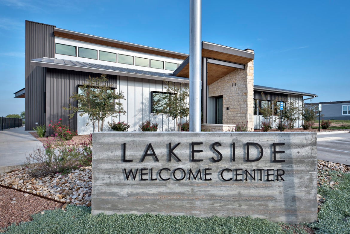 Modern building with large windows and a stone facade behind a concrete sign reading "Lakeside Welcome Center" on a landscaped property.