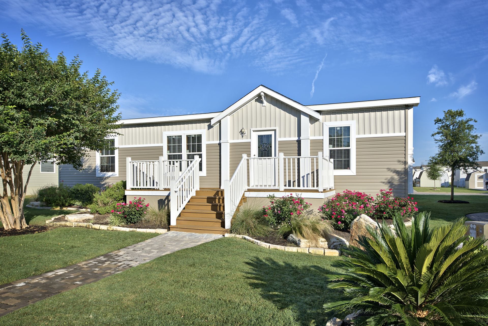 Single-story manufactured home with beige siding, white trim, front porch, stairs, landscaped yard, and a stone walkway under a blue sky.