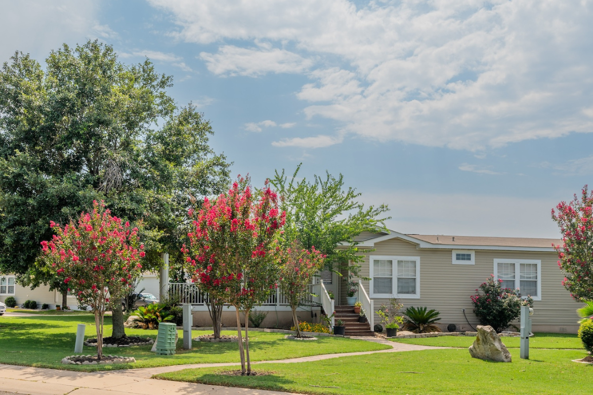 A beige manufactured home with a front porch, surrounded by green lawn, flowering trees, shrubs, and a large rock under a partly cloudy sky.