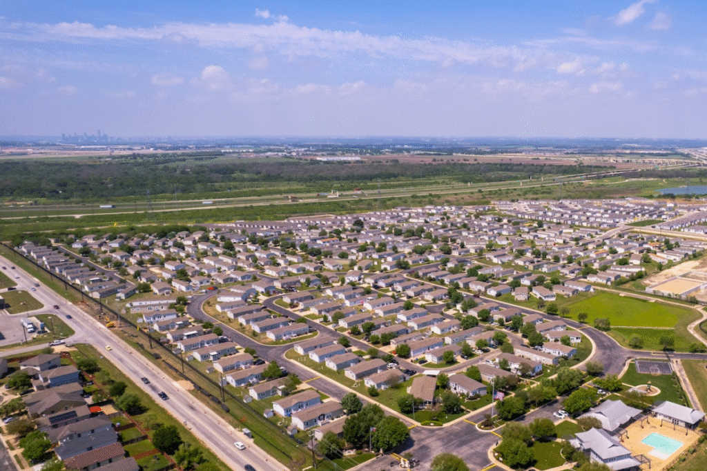 Aerial view of a suburban neighborhood with rows of houses, streets, and some greenery under a partly cloudy sky. City skyline visible in the distant background.