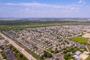 Aerial view of a suburban neighborhood with rows of houses, streets, and some greenery under a partly cloudy sky. City skyline visible in the distant background.